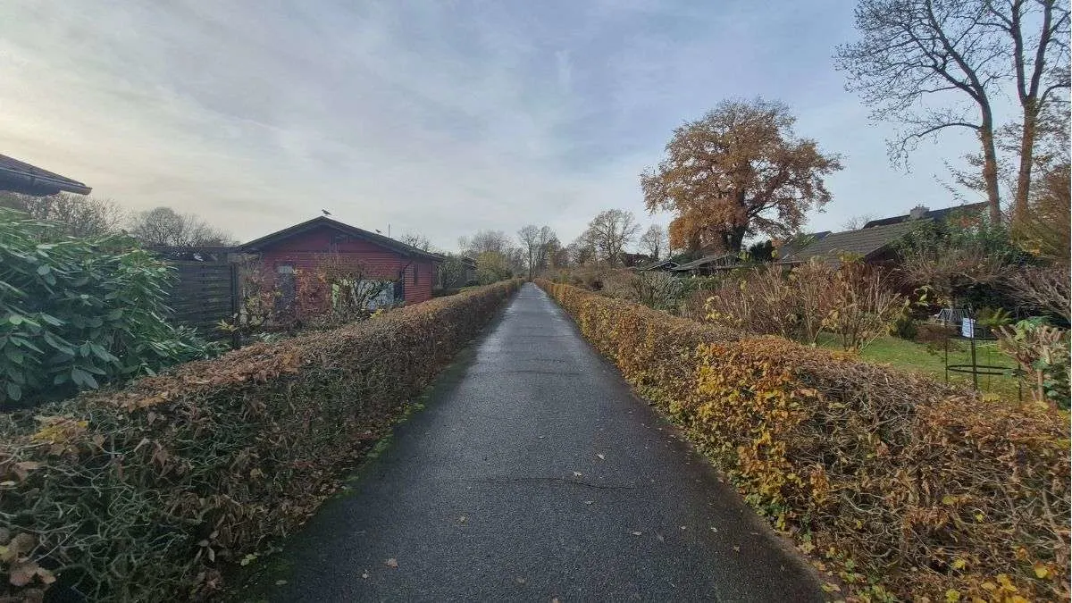 Ein asphaltierter Weg führt durch eine grüne Anlage, gesäumt von Hecken und Laubbäumen unter einem bewölkten Himmel. Holzhäuser stehen seitlich.