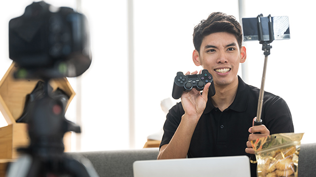 Ein Mann hält einen Controller und ein Telefon am Selfie-Stick. Eine Kamera steht auf einem Stativ. Ein Laptop ist sichtbar.