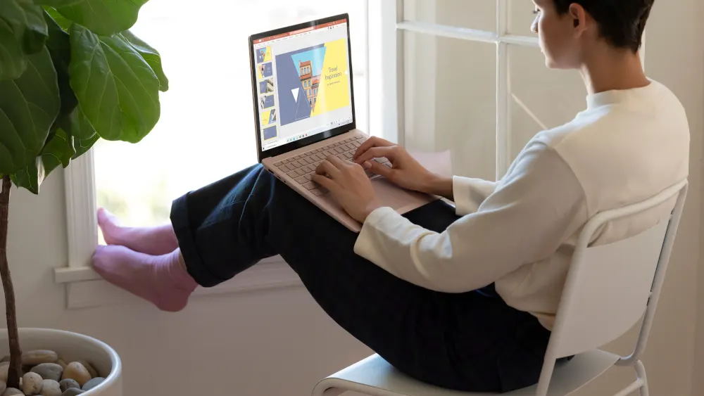 Eine Person sitzt mit einem Laptop am Fenster, die Füße hochgelegt. Ein Topf mit einer Pflanze steht links.