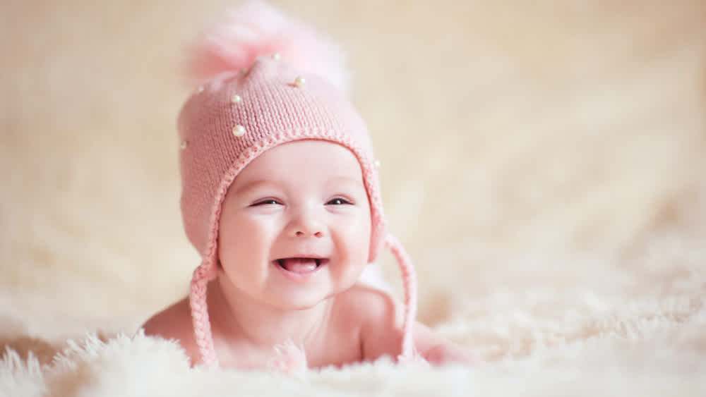 Ein Baby mit pinker Mütze liegt auf einer Decke und lacht.