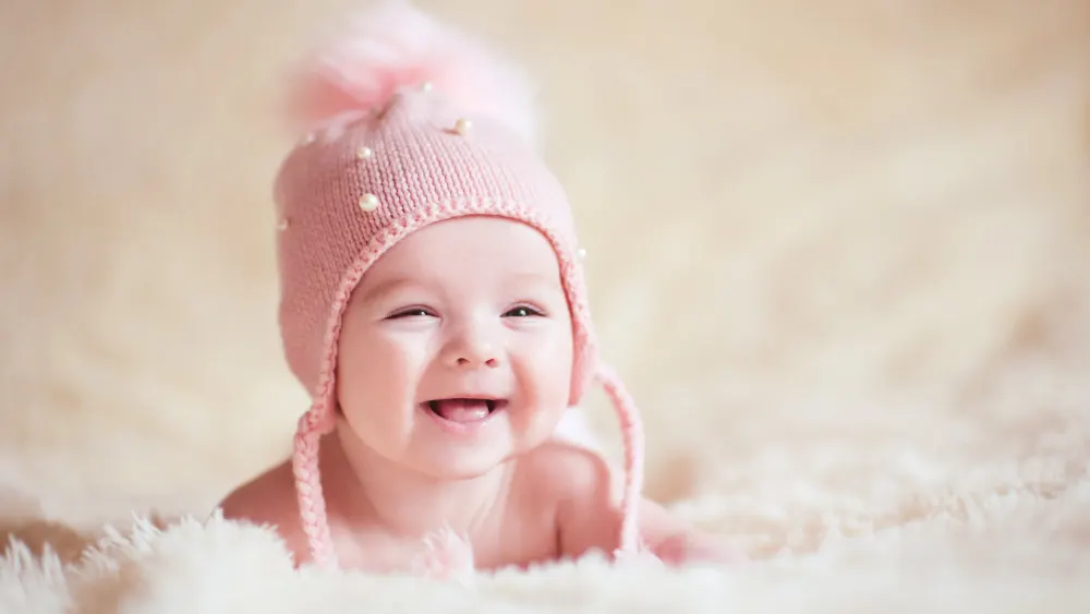 Ein lächelndes Baby liegt auf einer flauschigen Decke und trägt eine rosafarbene Mütze mit Bommel und Perlen.