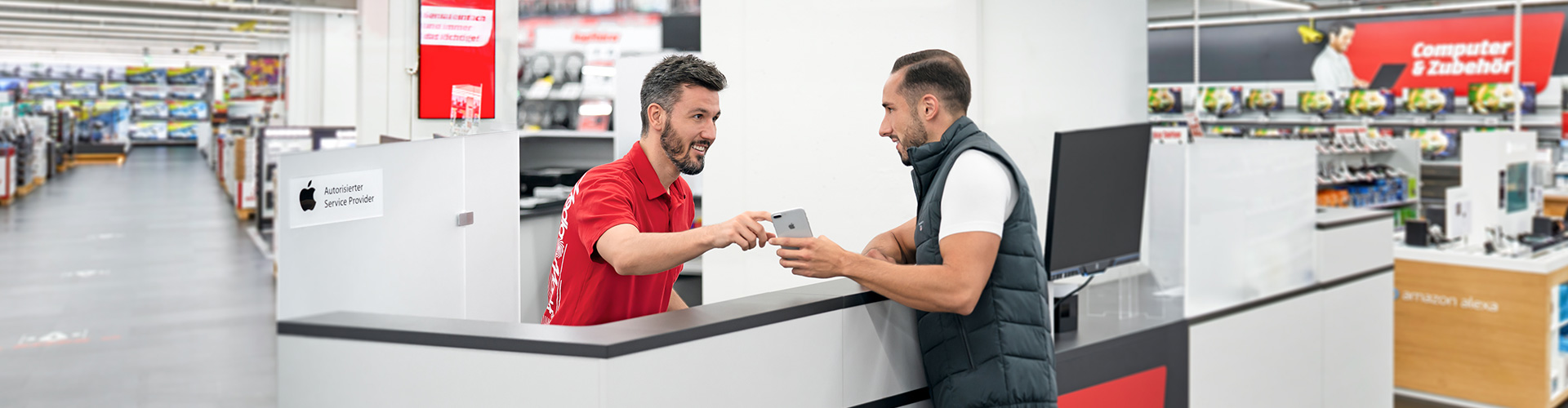 Ein Mann in rotem Hemd übergibt ein Telefon an einen Mann in Weste an einer grauen Theke in einem Geschäft.