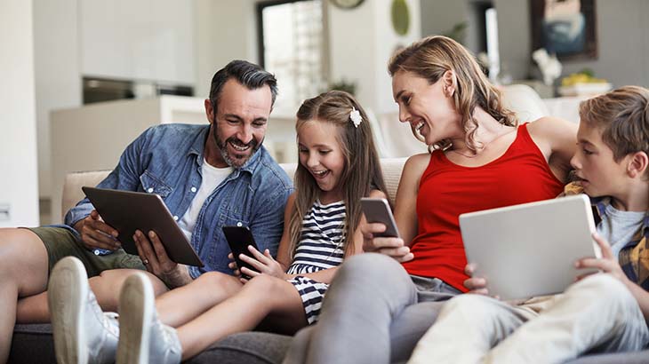 Eine Familie sitzt auf einem Sofa und benutzt Tablets und Handys. Sie tragen lässige Kleidung und scheinen sich zu amüsieren.