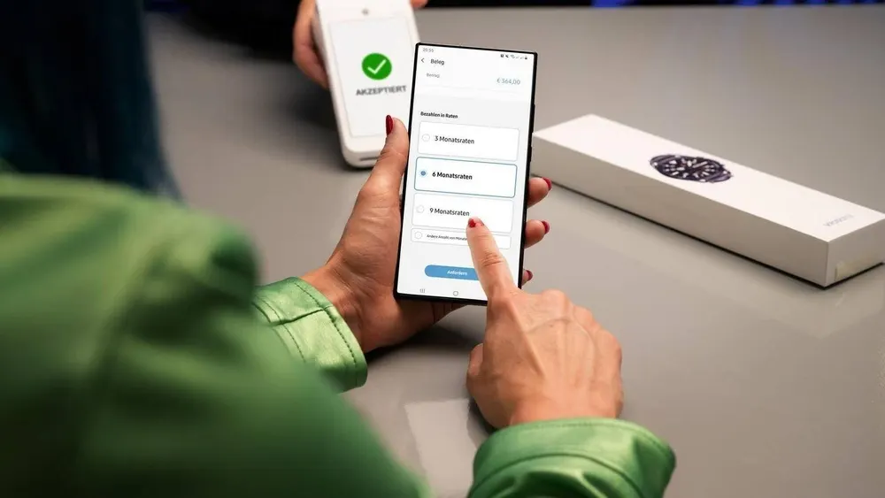 Eine Person hält ein Smartphone mit Ratenzahlungsoptionen in der Hand, neben einer weißen Uhrenverpackung und einem Zahlungsgerät mit grünem Haken.