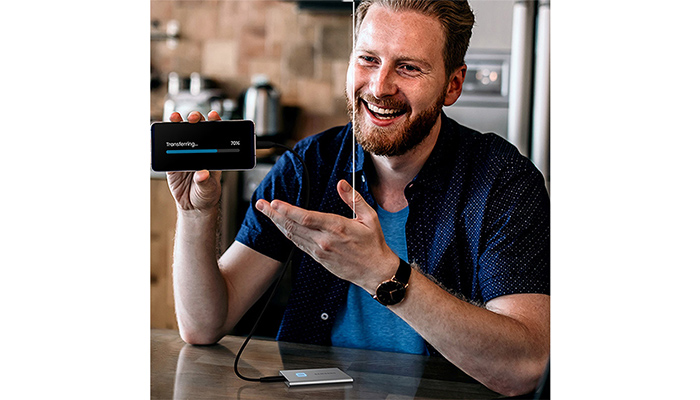 Ein Mann hält ein Handy mit einem Transferbildschirm. Eine externe Festplatte ist mit einem Kabel verbunden. Küche im Hintergrund.