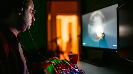 Ein Mann mit Headset und Brille tippt auf eine farbig beleuchtete Tastatur vor einem Monitor in einem dunklen Raum.