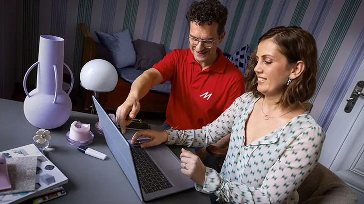 Zwei Personen arbeiten an einem Laptop an einem Tisch. Dekorative Gegenstände stehen auf dem Tisch. Eine Tür ist im Hintergrund.