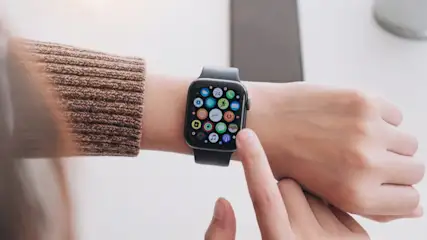 Eine Person mit braunem Pullover bedient eine Smartwatch mit bunten App-Symbolen am Handgelenk.