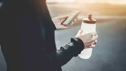 Eine Person hält ein Smartphone und eine weiße Wasserflasche mit braunem Deckel vor einem unscharfen Hintergrund.