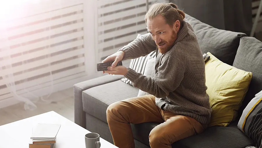 Ein Mann sitzt auf einem grauen Sofa und hält eine Fernbedienung in seinen Händen. Auf dem Tisch stehen Bücher und eine Tasse.