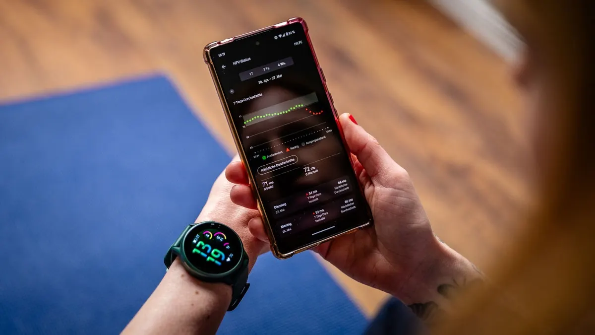 Eine Person hält ein Smartphone mit einer Gesundheits-App und trägt eine Smartwatch mit Gesundheitsdaten. Im Hintergrund ist eine blaue Yogamatte.