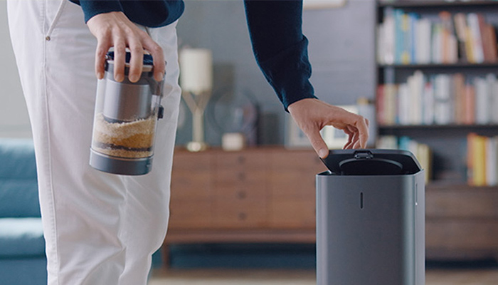 Eine Person leert einen Staubsaugerbehälter in einen grauen Abfallbehälter, im Hintergrund ein Bücherregal.