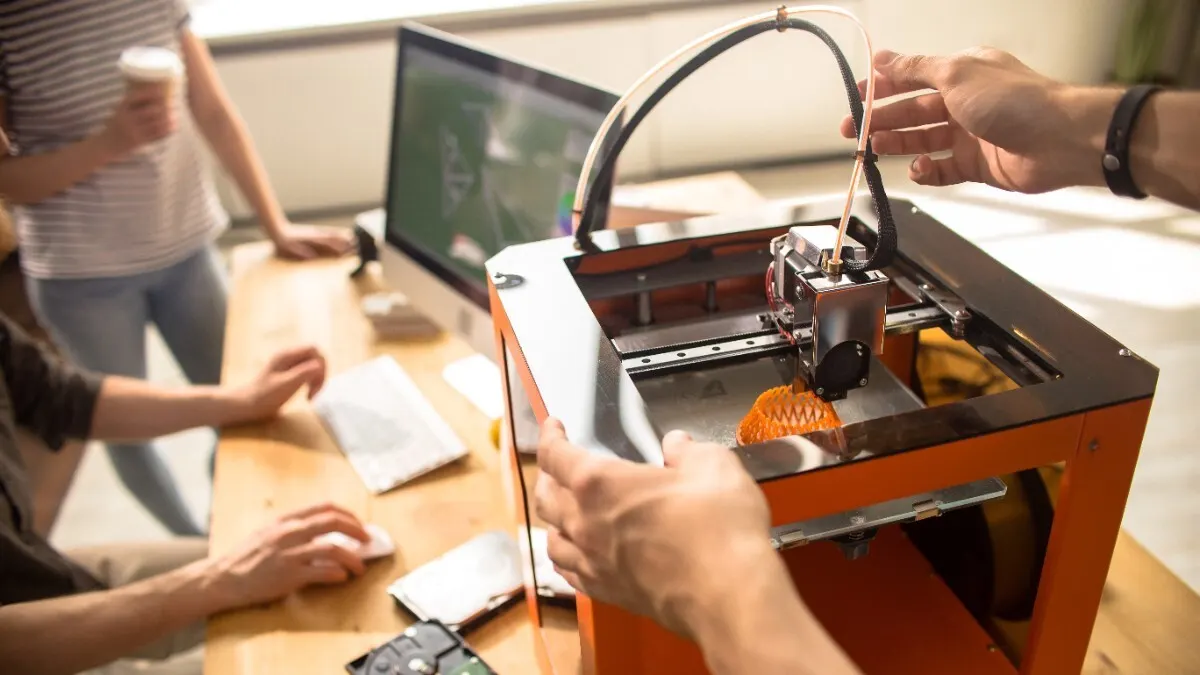 Ein 3D-Drucker steht auf einem Schreibtisch, darum herum stehen mehrere Personen, deren Hände zu sehen sind.
