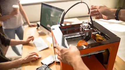 Ein 3D-Drucker steht auf einem Schreibtisch, darum herum stehen mehrere Personen, deren Hände zu sehen sind.