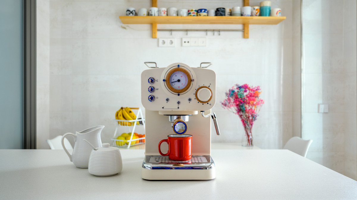 Eine helle Siebträgermaschine auf einem weißen Tisch, auf der Abtropfplatte des Geräts steht eine rote Tasse.