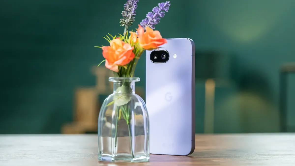 Ein violettes Smartphone steht neben einer Vase mit orangefarbenen Rosen und lila Lavendel auf einem Holztisch vor einer grünen Wand.