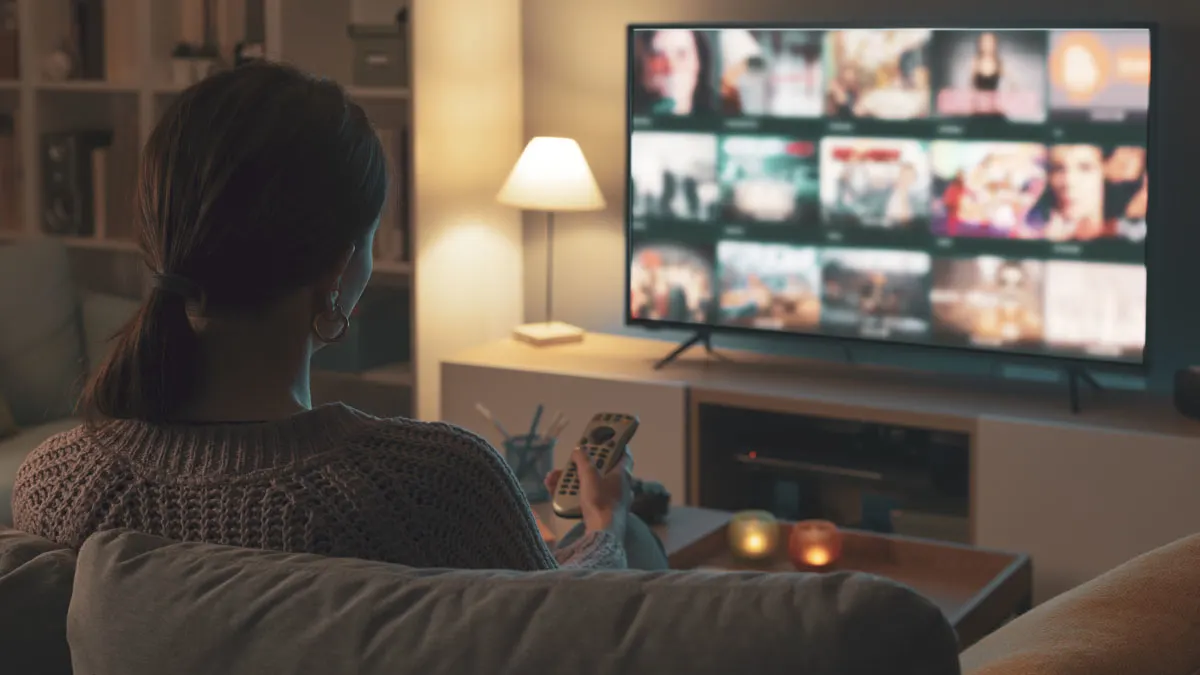 Eine Frau sitzt auf einem Sofa und hält eine Fernbedienung vor einem Fernseher, der mehrere Videos anzeigt.