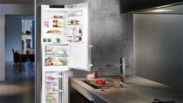 Offener weißer Kühlschrank voll mit Essen, neben einer Küchenspüle mit Obst und Armatur. Im Hintergrund ist ein Esstisch sichtbar.