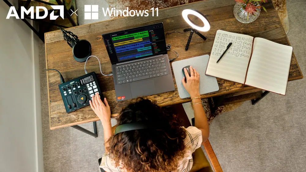 Draufsicht: Person sitzt am Holztisch mit Laptop, Audiomixer, Notizbuch, Lampe. AMD- und Windows 11-Logos sind sichtbar.