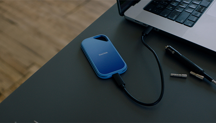 Blaue SanDisk Portable SSD per USB‑C an Laptop angeschlossen, liegt auf dunklem Tisch; daneben kleiner USB‑Stick.