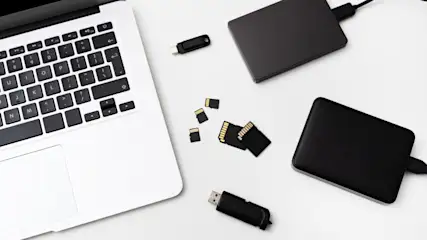 Ein Laptop mit schwarzer Tastatur liegt neben schwarzen Festplatten, USB-Sticks und SD-Karten auf einem weißen Tisch.