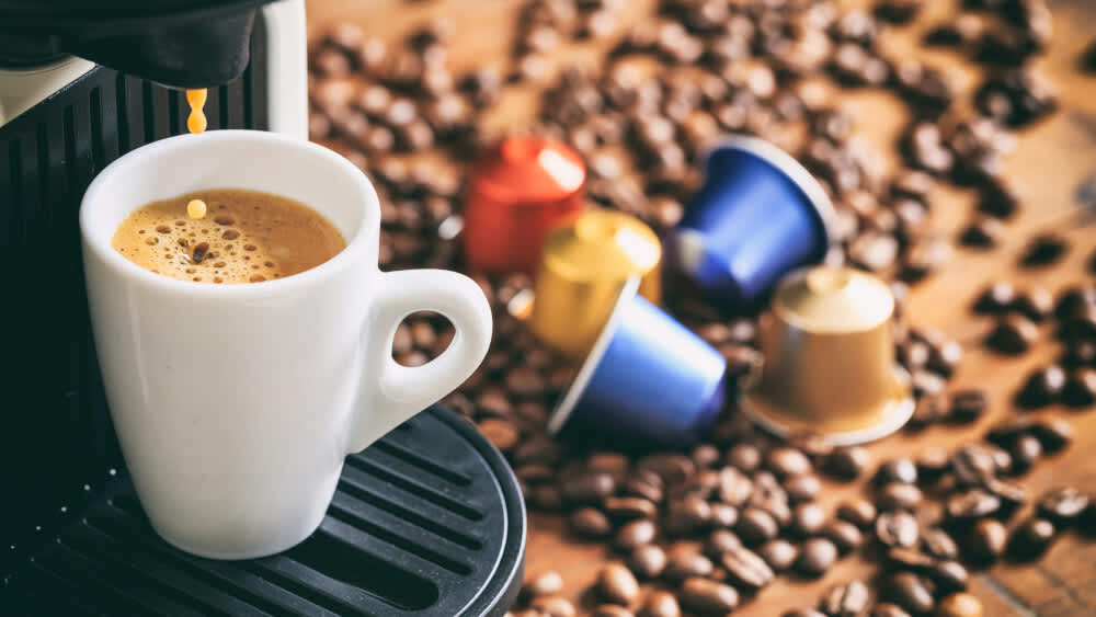 Mehrere Kaffee-Kapseln und Kaffeebohnen liegen neben einer Kaffeemaschine.