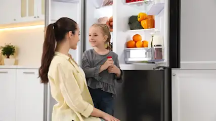 Eine Frau und ein Kind stehen vor einem offenen Kühlschrank in einer hellen Küche. Im Kühlschrank sind Orangen, Paprika und andere Lebensmittel.