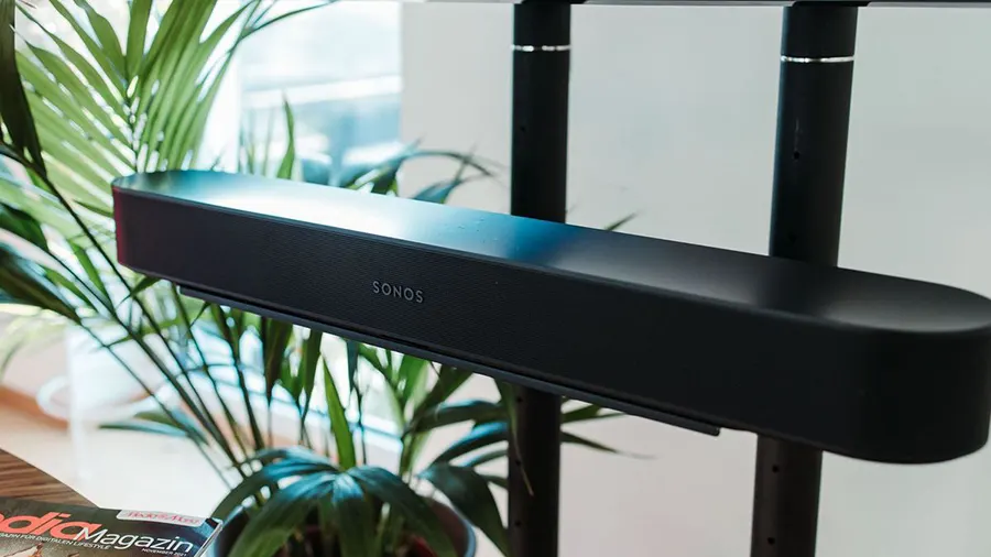 Schwarze Sonos Soundbar an Halterung vor grüner Pflanze und Magazin auf Tisch. Im Hintergrund sind schwarze Stangen.