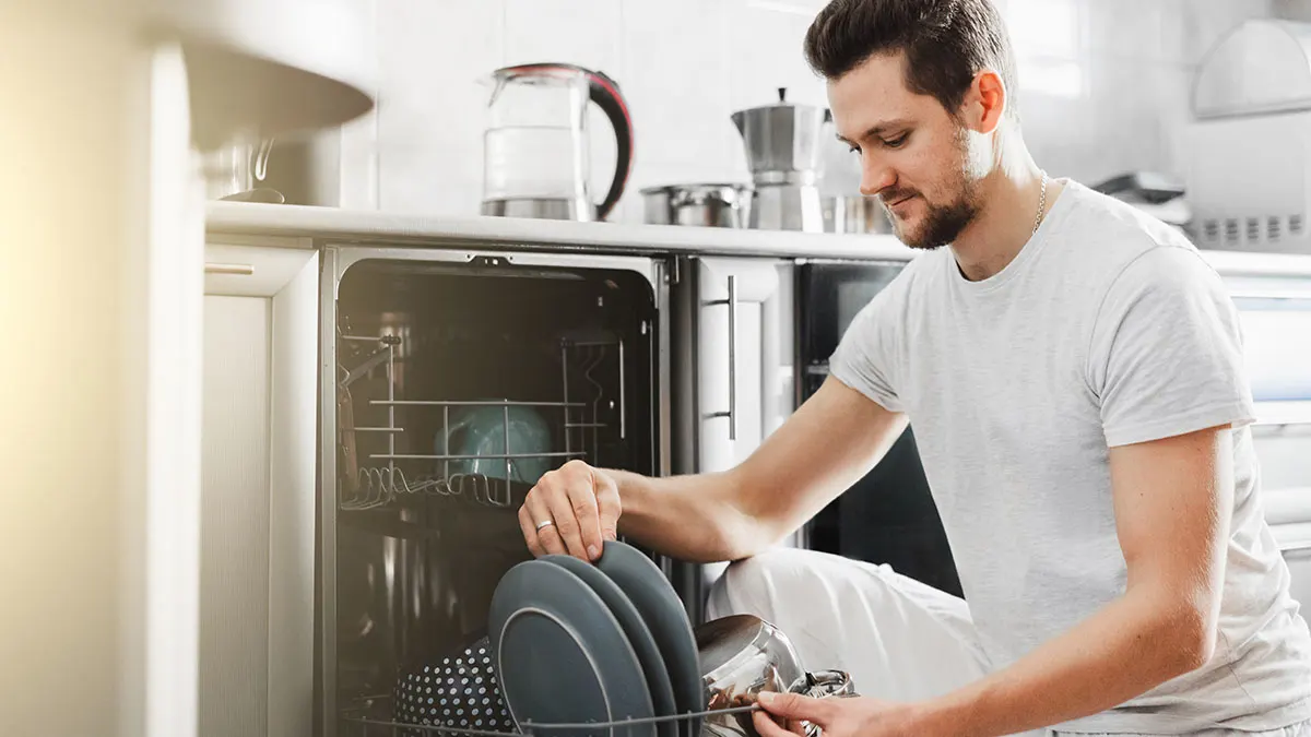 Mann im weißen T-Shirt belädt den unteren Korb eines Geschirrspülers mit Tellern und einem Topf.