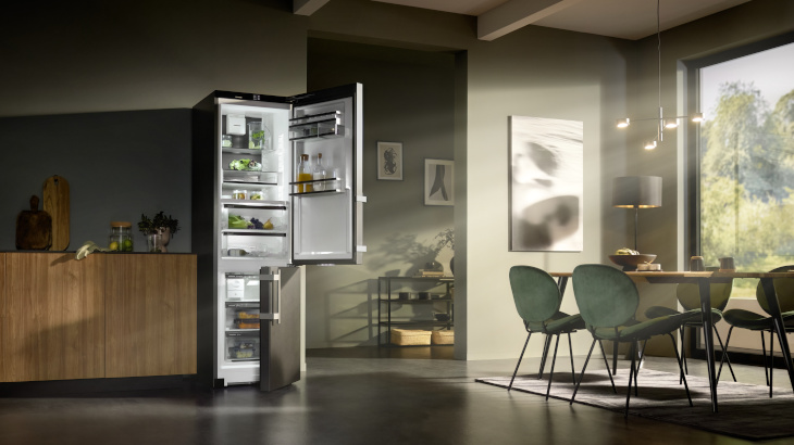 Ein offener Kühlschrank steht in einem Esszimmer mit Holzboden, Tisch, Stühlen und einem Fenster mit Blick auf Bäume.