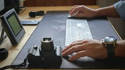 Schreibtisch mit Kamera, Tastatur, Uhr und digitalem Gerät. Hände bedienen Maus. Telefon und Armband liegen im Hintergrund.