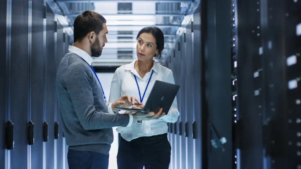 Zwei Personen in einem Serverraum. Eine Person hält einen Laptop. Serverschränke sind im Hintergrund sichtbar.