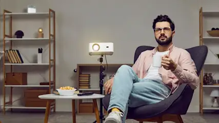 Ein Mann mit Brille sitzt in einem Sessel und hält eine Tasse. Neben ihm steht ein Projektor und ein Tisch mit Popcorn.