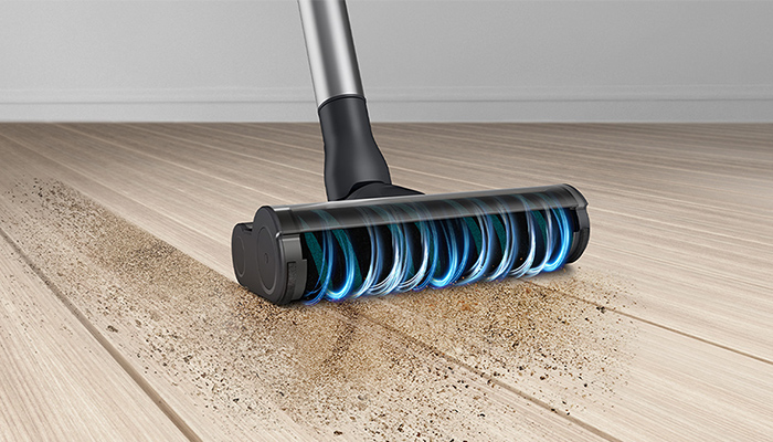 Ein Staubsauger mit Bürste reinigt einen hellen Holzboden mit Schmutz. Der Hintergrund ist eine helle Wand.