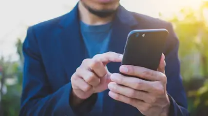 Ein Mann im blauen Jackett bedient ein schwarzes Smartphone. Der Hintergrund ist unscharf und zeigt grüne Vegetation.