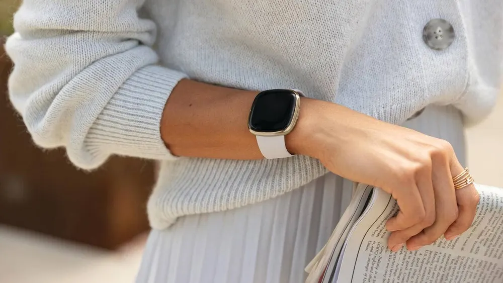 Eine Person trägt eine Smartwatch mit weißem Armband und einen hellen Strickpullover und hält eine Zeitung.
