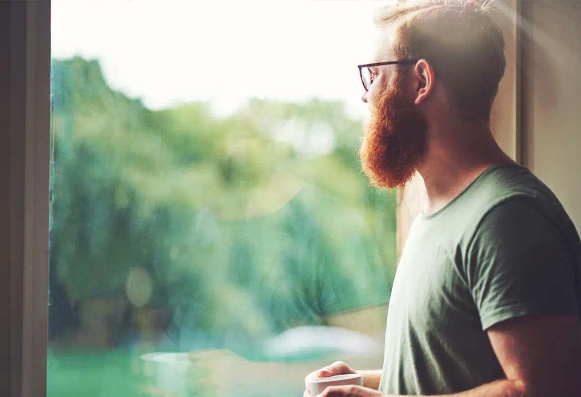 Ein Mann mit Brille und T-Shirt blickt aus dem Fenster. Er hält eine Tasse. Draußen sind Bäume und Grün zu sehen.