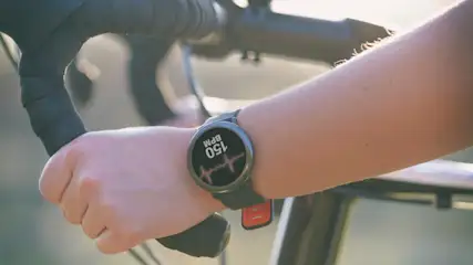 Nahaufnahme einer Hand am Fahrradlenker, eine Smartwatch zeigt einen Puls von 150 BPM an.