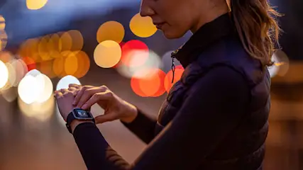 Frau im dunklen Oberteil blickt auf eine Smartwatch vor verschwommenen Lichtern.