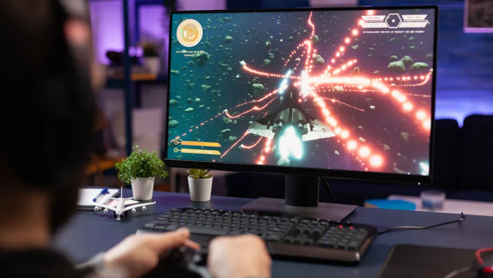 Eine Person spielt ein Weltraum-Videospiel auf einem Computer mit einer Tastatur, zwei Topfpflanzen und einem Flugzeugmodell auf dem Tisch.