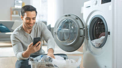Ein Mann hält ein Smartphone in der Hand und sitzt neben einer Waschmaschine.