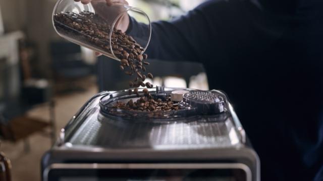 Kaffeebohnen werden aus einem Glasbehälter in eine silberne Kaffeemühle gegossen.