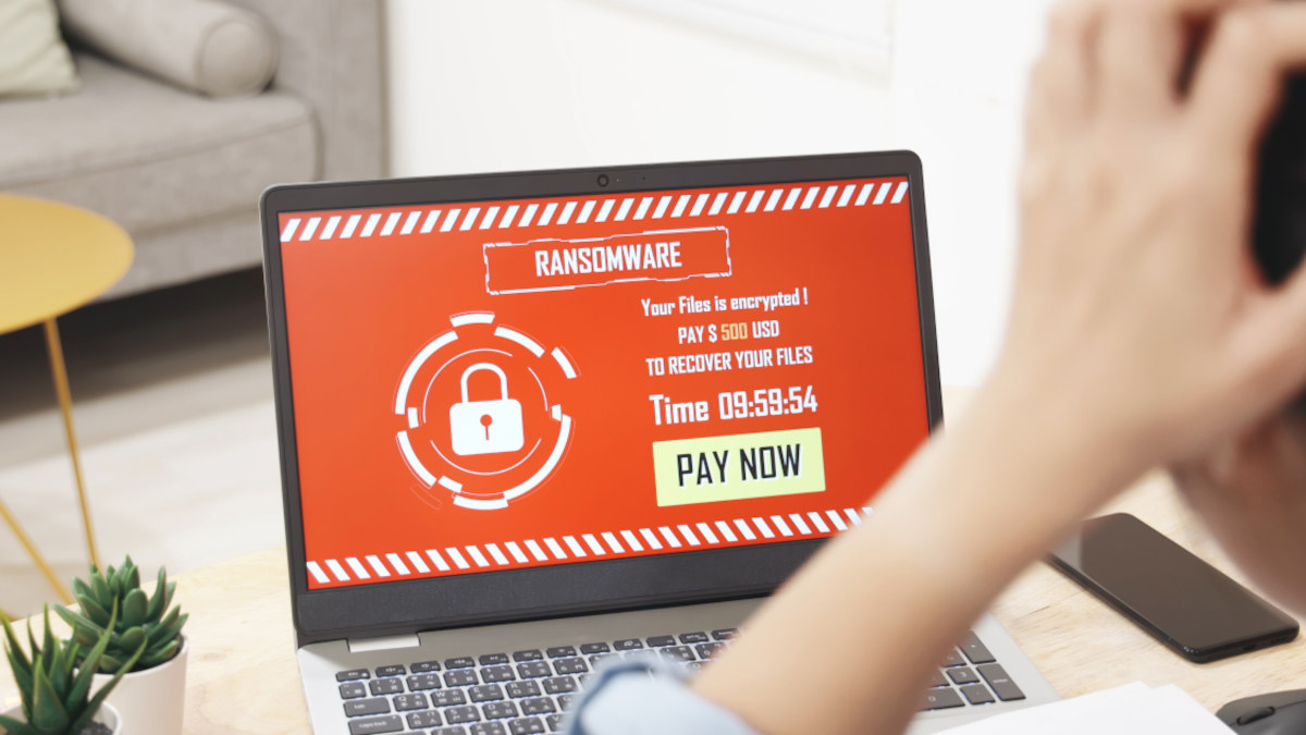 Laptop-Bildschirm zeigt rote Ransomware-Warnung mit weißem Schloss-Symbol, Text fordert Zahlung von 500 USD. Person hält sich den Kopf.