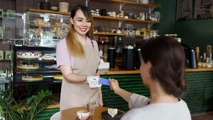 Kellnerin in einem Café hält ein mobiles Kartenterminal, während eine sitzende Kundin mit einer kontaktlosen Karte bezahlt; auf dem Tisch stehen Kaffee und Kuchen, im Hintergrund sind Theke und Kuchenvitrine zu sehen.
