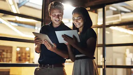 Ein Mann und eine Frau stehen vor einer Glaswand. Beide halten Tablets und lächeln.