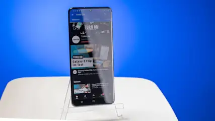 Ein schwarzes Smartphone auf einem transparenten Ständer, das eine Webseite mit dem Titel "TURN ON" anzeigt, vor einem blauen Hintergrund.