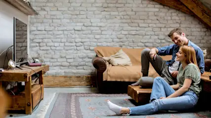 Ein Paar entspannt im Wohnzimmer mit Ziegelwand, Fernseher auf einem Holztisch, Teppich auf dem Boden und braunem Sofa.