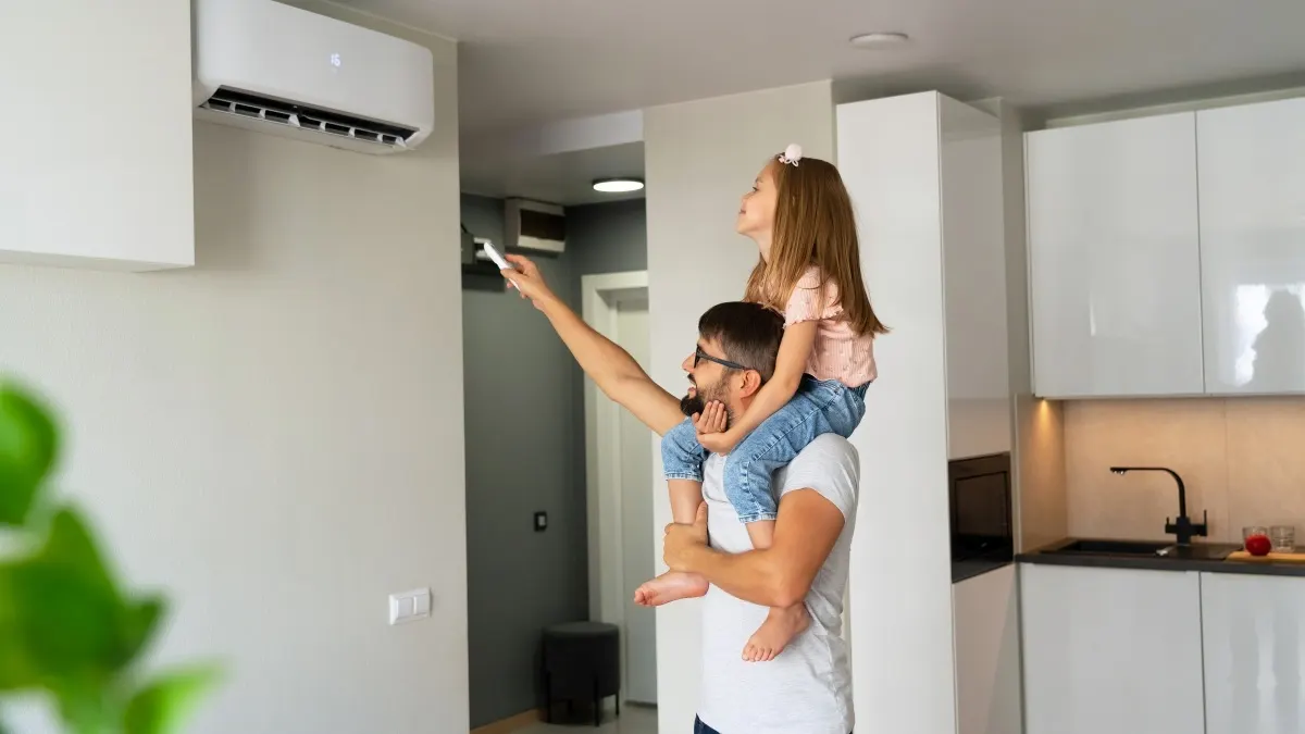 Ein Mann trägt ein Mädchen auf den Schultern und bedient per Fernbedienung eine an der Wand montierte Klimaanlage.