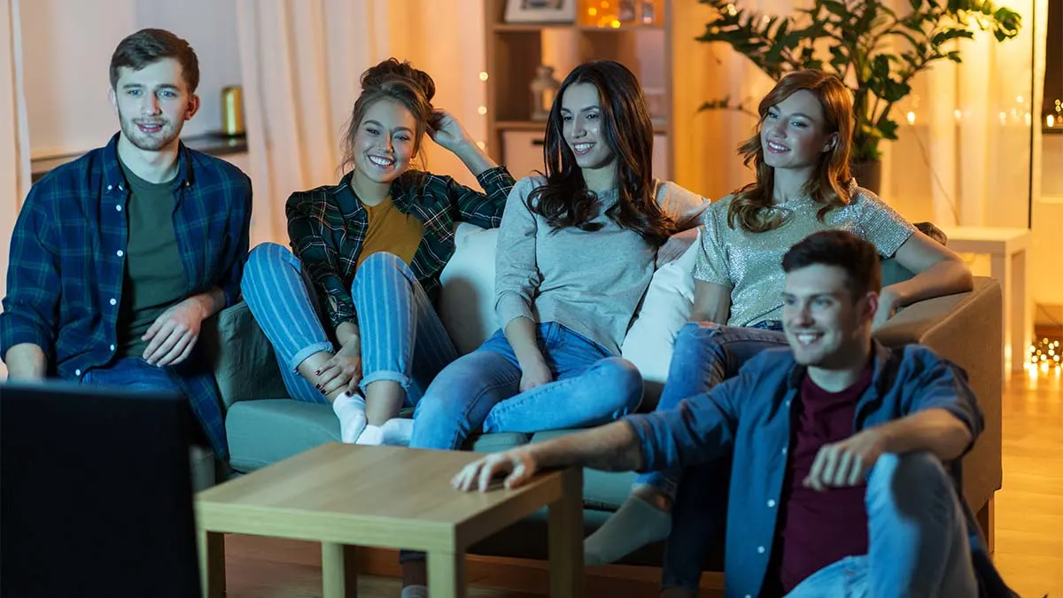 Fünf junge Leute sitzen auf einem Sofa und schauen lächelnd auf einen Fernseher im Wohnzimmer.