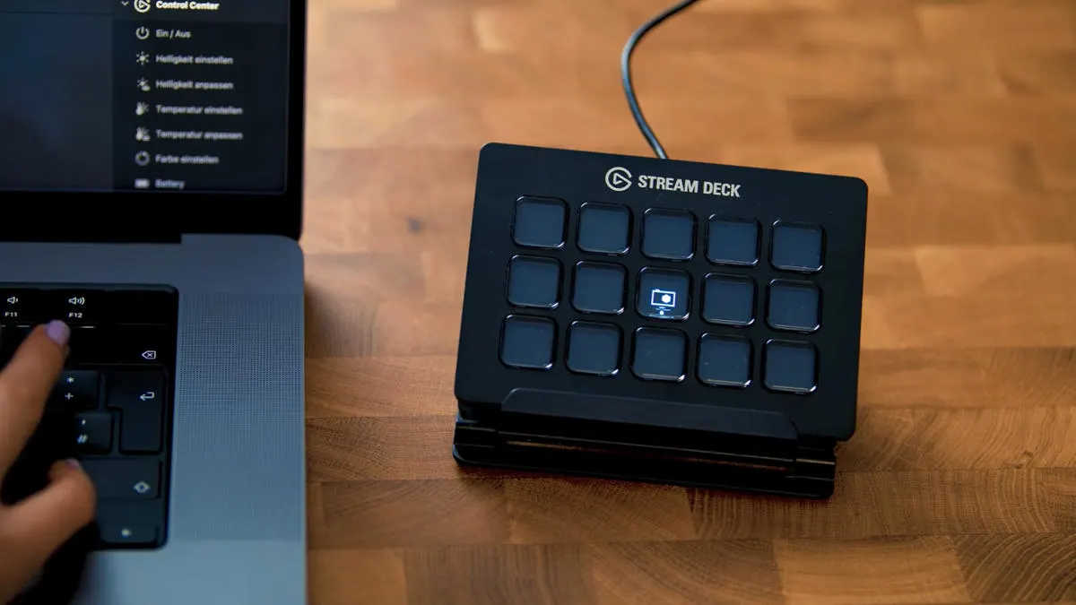 Das Stream Deck von Elgato steht auf einem Holztisch neben einem Laptop, auf dem eine Hand tippt.
